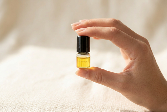 Hand holding a single small Lazaval Discovery Set perfume oil vial with a black cap, showing the vial's small scale against the fingers, golden natural oil inside, in soft daylight.
