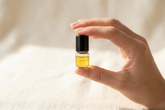 Hand holding a single small Lazaval Discovery Set perfume oil vial with a black cap, showing the vial's small scale against the fingers, golden natural oil inside, in soft daylight.